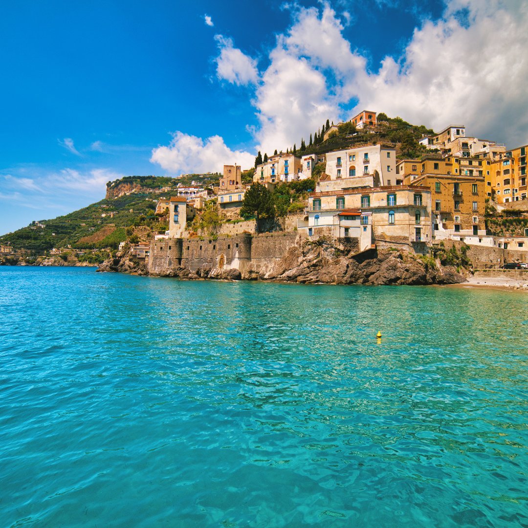 A pastel Amalfi Coast village seen from the sea, with houses stacked above clear turquoise water and a steep headland