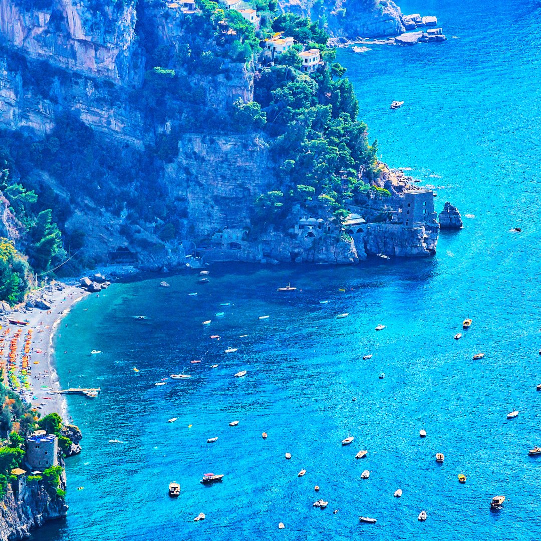 An aerial view of a sheltered cove on the Amalfi Coast with small fishing boats anchored in deep blue water and a pebble beach below limestone cliffs
