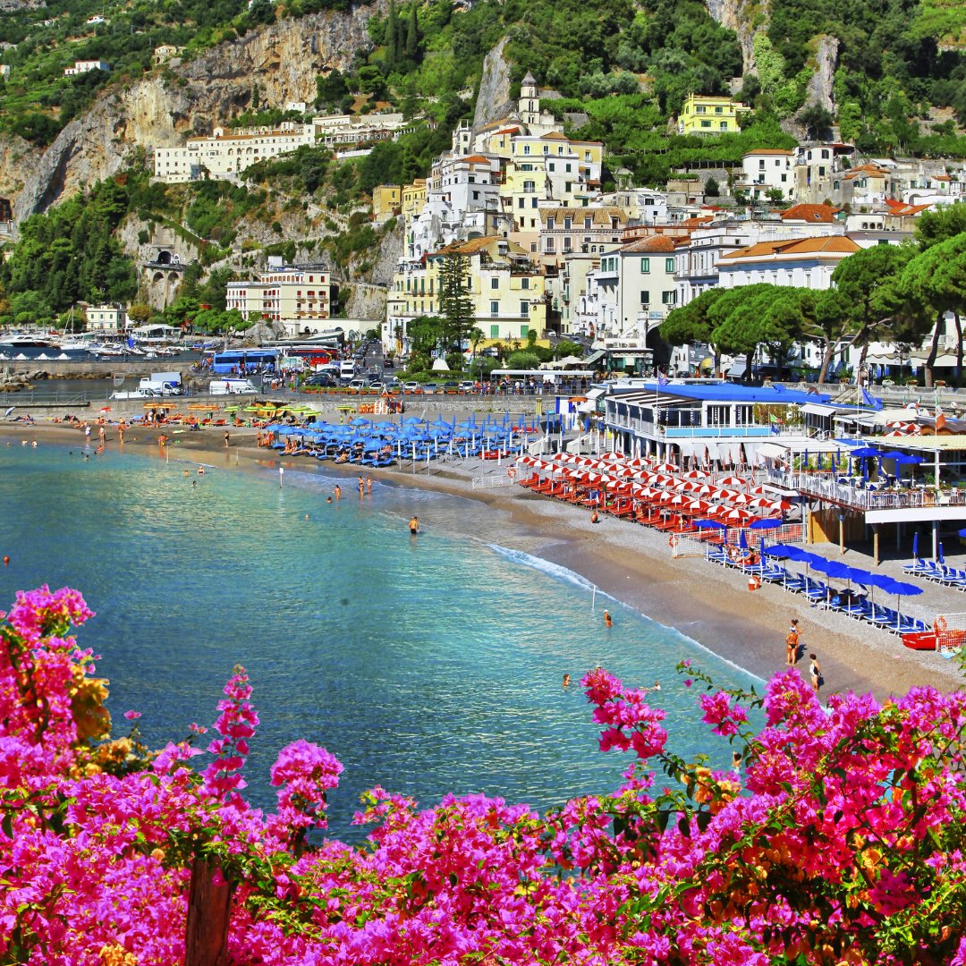 Amalfi's main beach in summer with bougainvillea in the foreground, striped beach umbrellas in red and blue, and the medieval town climbing the hillside