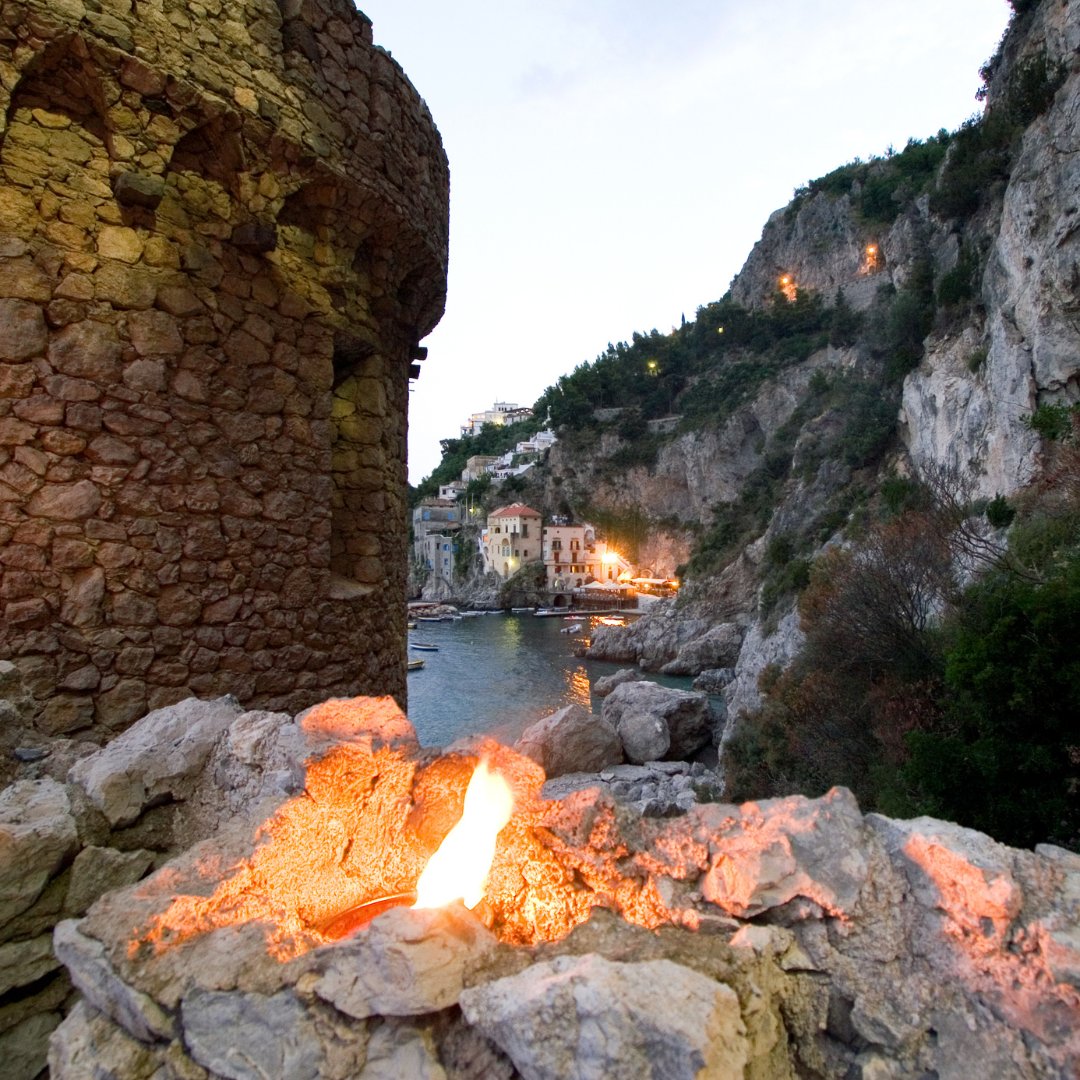 A medieval Saracen watchtower at dusk above a sheltered cove on the Amalfi Coast, with a fire burning in the foreground