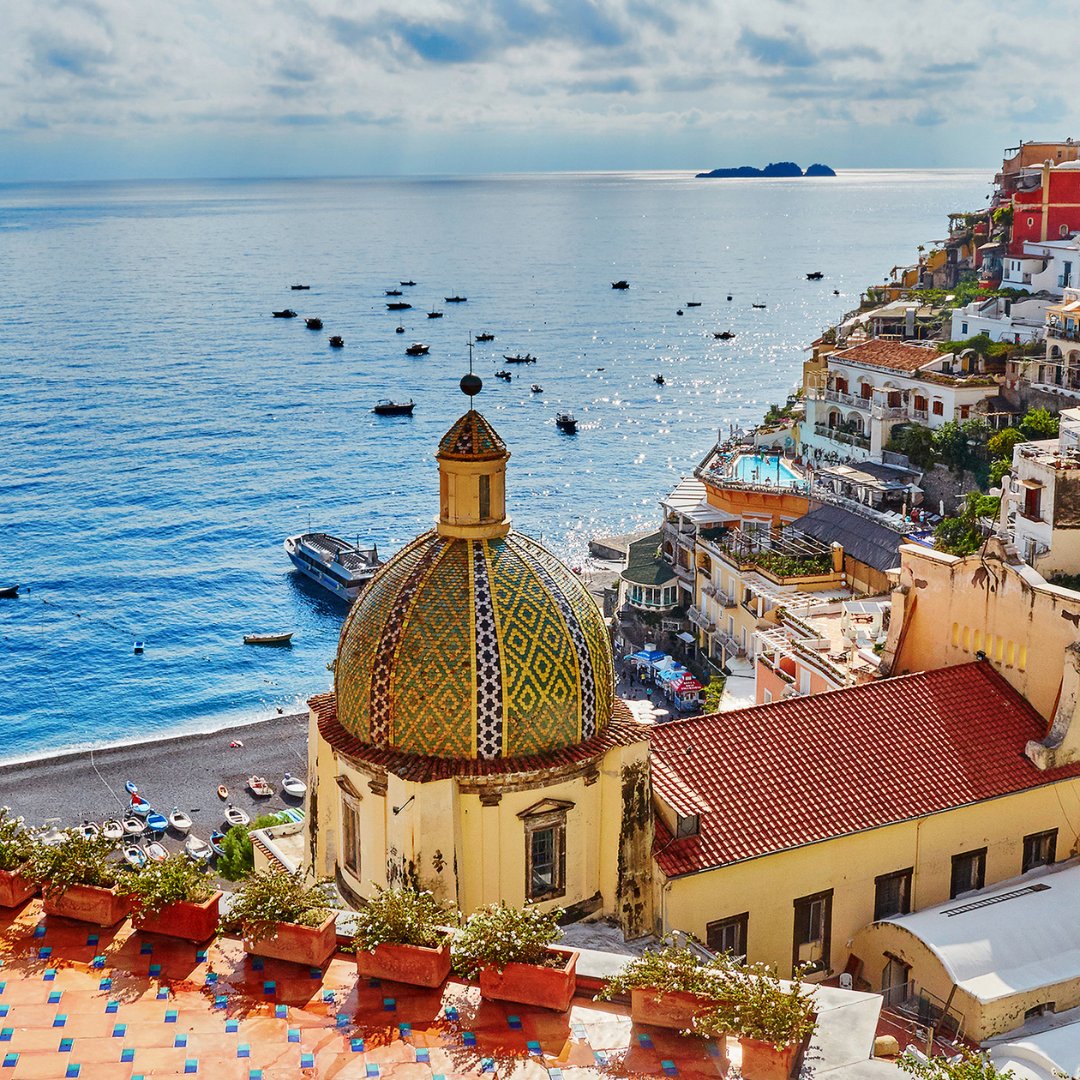 The majolica-tiled dome of Santa Maria Assunta in Positano with terracotta rooftops descending to the beach and fishing boats on the Tyrrhenian Sea