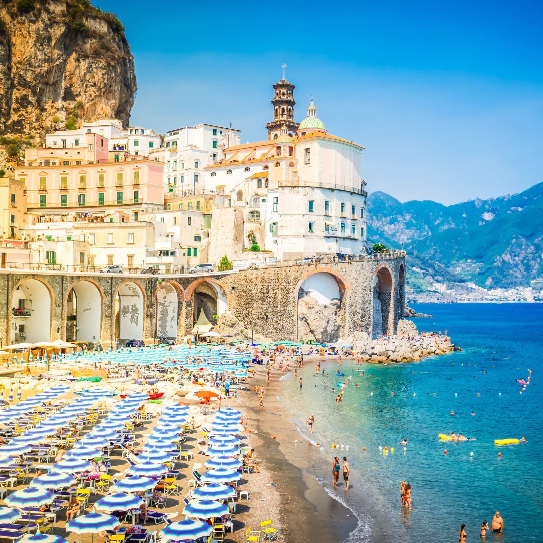 Atrani beach in summer with rows of umbrellas, bathers in the shallows and the cliff-top bell tower of the Collegiata di Santa Maria Maddalena