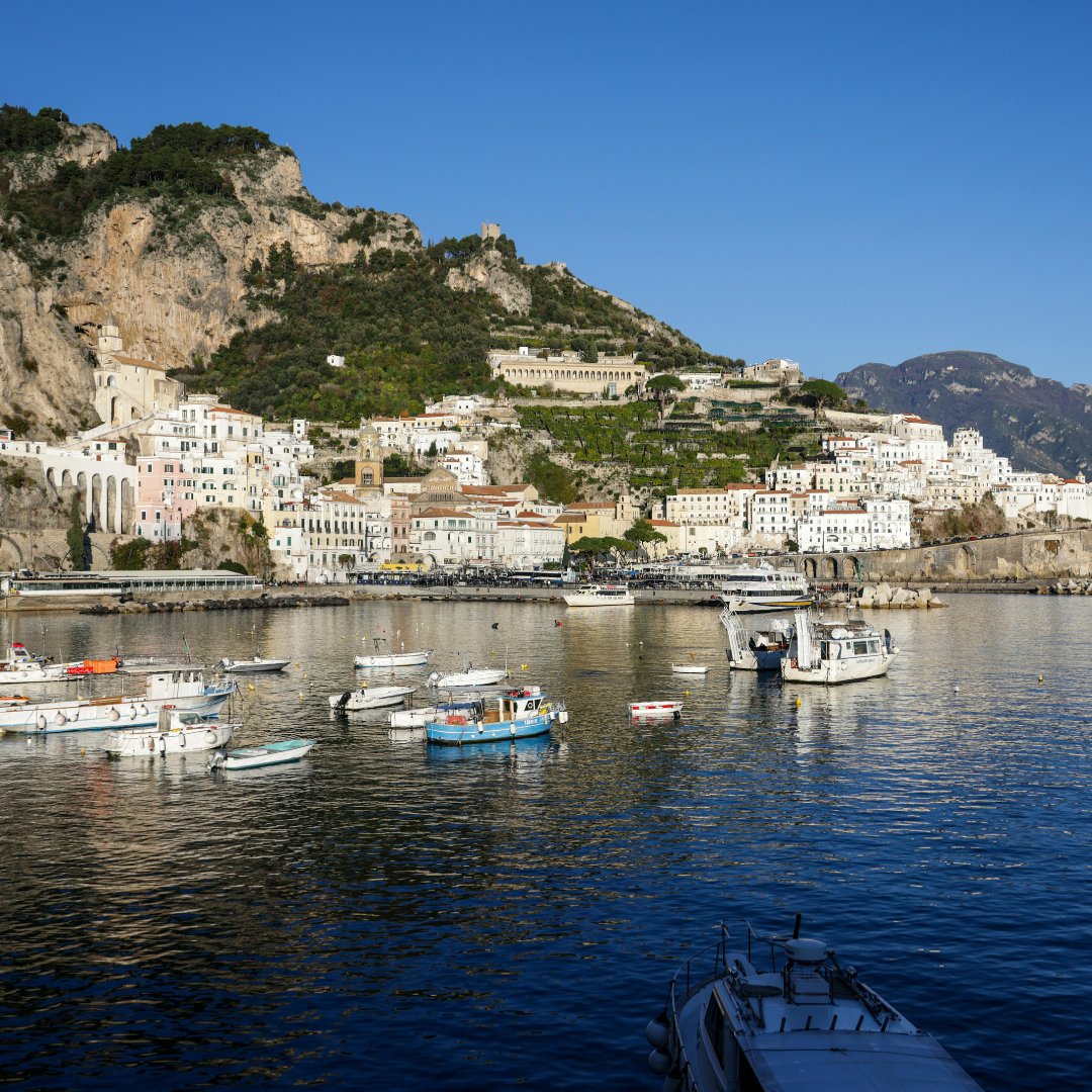 Amalfi harbour at morning light with fishing boats moored along the breakwater and the medieval town climbing the hillside behind, Campania, southern Italy